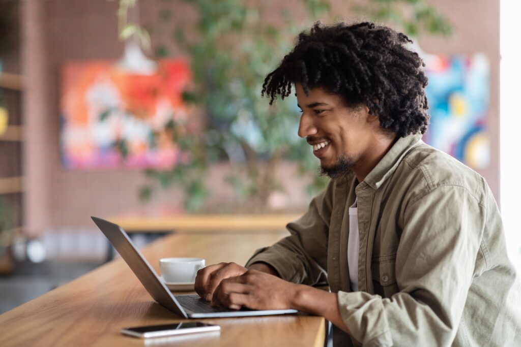 stock-photo-cheerful-young-african-american-curly-male-typing-on-laptop-at-table-with-coffee-cup-and-phone-in-2149804729