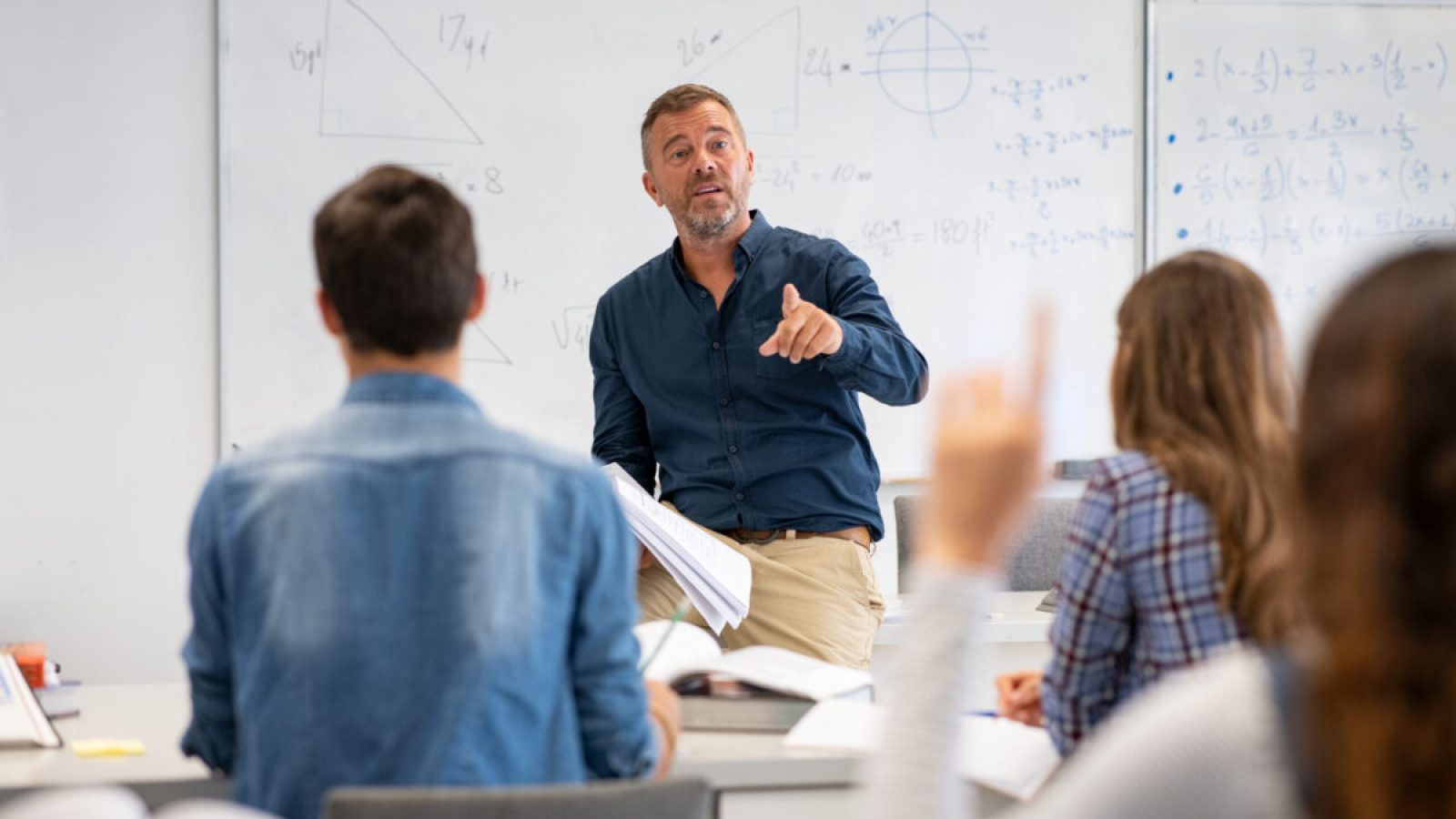 stock-photo-professor-pointing-at-college-student-with-hand-raised-in-classroom-student-raising-a-hand-with-a-1836272266